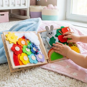 May include: A fabric quiet book featuring felt pages with interactive elements including a yellow sun, a white cloud, and a rainbow made of red, orange, yellow, green, blue, and purple flowers. The opposite page shows a gray felt rabbit.