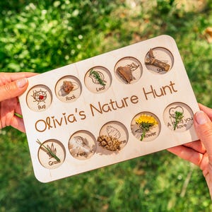 May include: A wooden nature hunt board with engraved circles containing various natural items like a bug, rock, leaf, and flower. The board is held by hands and features the text "Olivia's Nature Hunt".