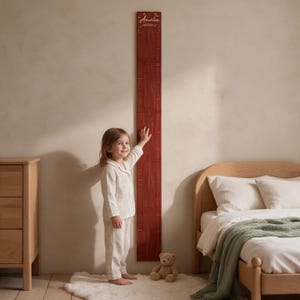 May include: A tall, vertical wooden growth chart with a dark reddish-brown finish, featuring engraved measurement markings and the name Amelia printed in white script at the top. The board is mounted on a wall, displaying a scale.