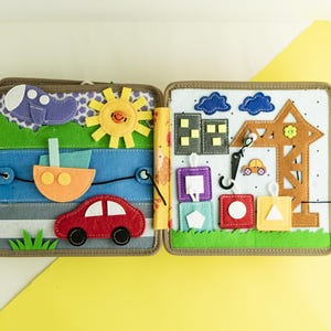 May include: A felt activity book for toddlers with a variety of textures and shapes. The book features a car, a plane, a boat, a sun, a crane, and a building. The book is designed to help children develop their fine motor skills and learn about different shapes and colors.