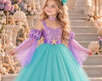 Puede incluir: Un vestido largo hasta el suelo con un corpiño morado de lentejuelas, una falda de tul turquesa voluminosa y mangas abullonadas moradas transparentes. El vestido incluye un fajín morado con un adorno de estrella de mar, gargantilla y lazo.