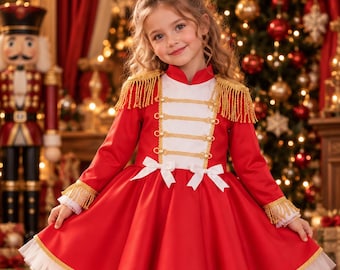 Puede incluir: Un vestido rojo y blanco con hombreras de flecos dorados, adornos trenzados dorados y lazos blancos en el corpiño. El vestido tiene mangas largas con puños de flecos dorados y una falda acampanada con un dobladillo de tul blanco.