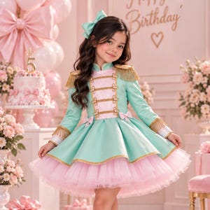 May include: A mint green and light pink dress with gold fringe epaulets, gold braided trim, and a layered tulle skirt. The dress features a high collar, long sleeves, and a matching mint green hair bow. The outfit is paired with light pink boots.