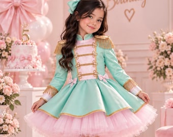 May include: A mint green and light pink dress with gold fringe epaulettes, gold braided trim, and a layered tulle skirt. The dress features a high collar, long sleeves, and a matching mint green hair bow. The outfit is paired with light pink boots.