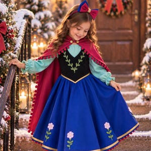May include: A dress featuring a black bodice with green leaf embroidery, long light blue sleeves, and a full blue skirt with pink flower appliqués. A deep red cape with red pom-pom trim and a matching red and blue hair bow complete the ensemble.