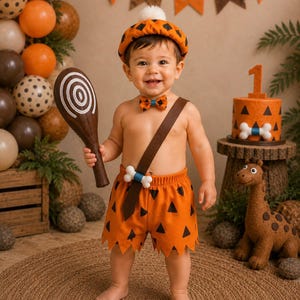 May include: A prehistoric-themed costume set consisting of orange shorts with black triangles, a matching bow tie, a brown sash with a white bone accent, and an orange beret with black triangles and a white pom-pom, paired with a brown club prop.