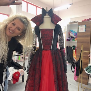 May include: A floor-length gown featuring a red bodice with a black lace overlay, long black lace sleeves, and a high standing collar. The skirt consists of a red base layer with a black lace overlay and a black bow at the neckline.