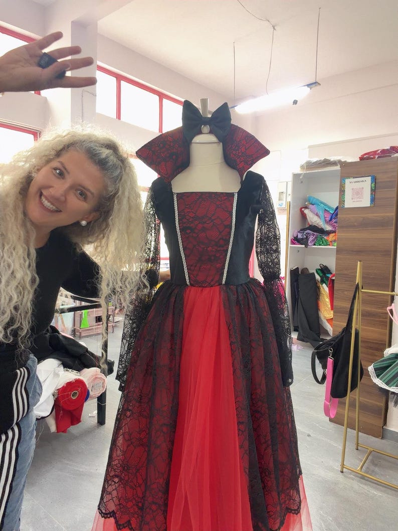 May include: A floor-length gown featuring a red bodice with a black lace overlay, long black lace sleeves, and a high standing collar. The skirt consists of a red base layer with a black lace overlay and a black bow at the neckline.