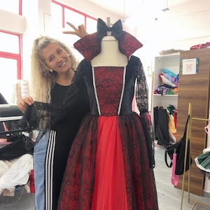 May include: A floor-length gown featuring a red bodice and skirt overlaid with black lace, long sheer black lace sleeves, a high standing collar, and a black bow at the neckline. The dress has a square neckline and a full, layered skirt.