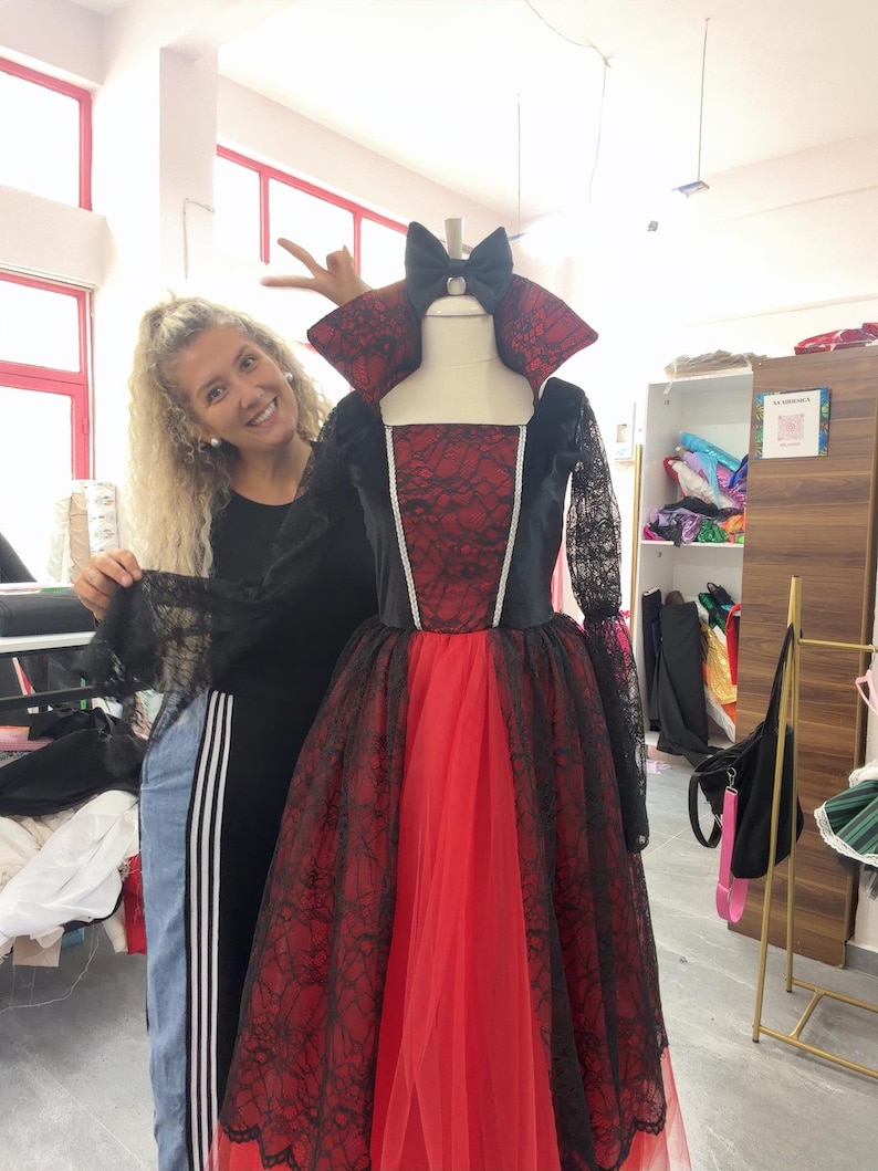 May include: A floor-length gown featuring a red bodice and skirt overlaid with black lace, long sheer black lace sleeves, a high standing collar, and a black bow at the neckline. The dress has a square neckline and a full, layered skirt.