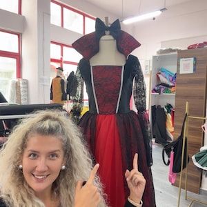 May include: A floor-length dress featuring a red bodice with a black lace overlay, a high standing collar, long black lace sleeves, and a full red tulle skirt. A black bow is attached at the neckline, and silver trim accents the bodice seams.