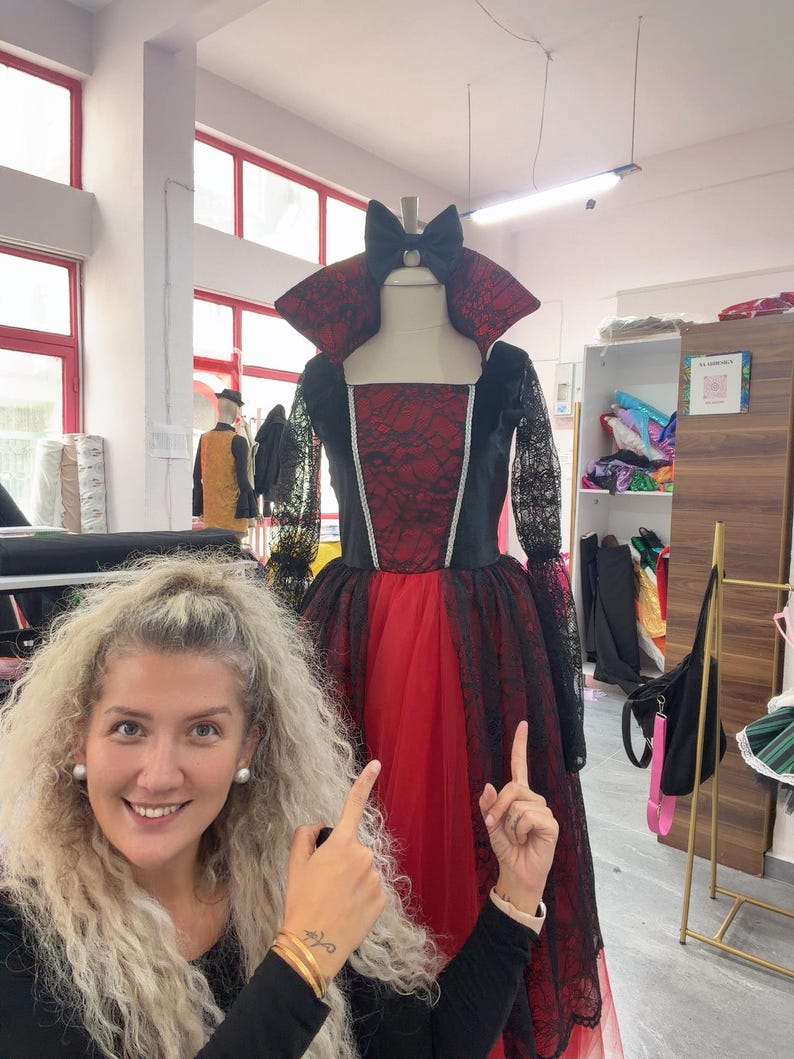 May include: A floor-length dress featuring a red bodice with a black lace overlay, a high standing collar, long black lace sleeves, and a full red tulle skirt. A black bow is attached at the neckline, and silver trim accents the bodice seams.