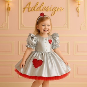 May include: A silver dress with short puffed sleeves, a round neckline, and a full skirt. The dress features red heart-shaped appliqués with white beaded borders on the bodice and skirt. It includes a red tulle hem and a matching red heart headband.