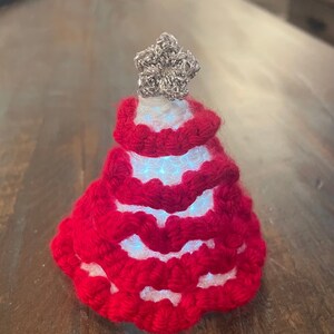 Può includere: Un albero di Natale fatto a mano, rosso, all'uncinetto, con una stella argentata in cima. L'albero ha un interno bianco che si illumina con piccole luci. L'albero è appoggiato su una superficie di legno scuro.