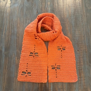 Op de afbeelding: Een gebreide sjaal in een felle oranje kleur met een herhalend patroon van kleine uitgesneden bloemen.
