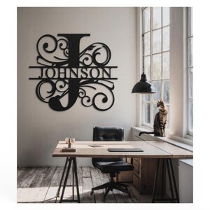 May include: Black metal wall decor featuring a large letter J and the word "JOHNSON" in a decorative font. The decor is hanging on a white wall in a room with a wooden floor, a desk, and a chair.