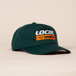 Pode incluir: Boné de beisebol verde escuro com a inscrição "LOCAL SPORTS" bordada em branco e laranja. O boné tem uma viseira curva e uma coroa estruturada. O texto é em negrito, sem serifa.