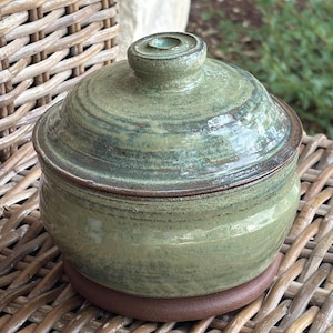 May include: A round ceramic jar with a lid, featuring a glossy sage green glaze with subtle brown undertones and a visible horizontal texture. The base of the jar is made of unglazed, smooth brown clay, and the lid has a small, centered knob handle.