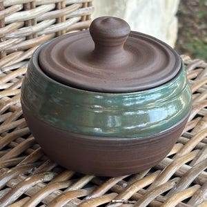 May include: A round, dark brown ceramic jar with a matching lid featuring a central knob. The upper portion of the jar is finished with a glossy, forest green glaze that contrasts with the matte, textured dark brown clay of the base and lid.