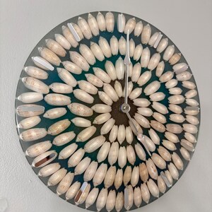 May include: A circular wall clock featuring a deep teal resin base embedded with numerous white, oval-shaped seashells arranged in concentric circles. The clock face includes silver-toned hour markers and thin, silver-colored metal clock hands.