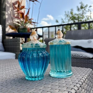 May include: Two glass jars with ribbed textures and vibrant blue hues. The left jar has a rounded, bulbous body, while the right jar is cylindrical. Both feature clear glass lids topped with decorative, swirling amber and white accents.