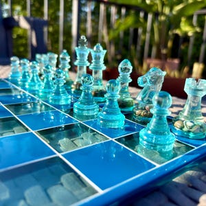 May include: A glass chess set with turquoise pieces on a blue and clear glass board. The pieces include pawns, rooks, knights, bishops, and a queen and king. The board is set on a light-colored surface with a blurred background.