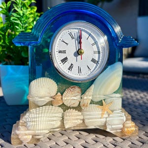 May include: A mantel clock encased in translucent blue resin with an arched top. The base is filled with embedded seashells, a small starfish, and sand. The clock face is white with black Roman numerals and thin black hands with a red second hand.