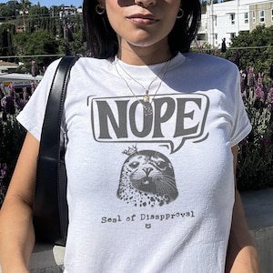 Może przedstawiać: Biała koszulka z nadrukiem przedstawiającym fokę w koronie i dymkiem z napisem "NOPE". Pod nadrukiem znajduje się tekst "Seal of Disapproval".