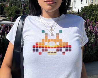 May include: White t-shirt with a pixelated graphic of a spaceship with a sunset design. The spaceship is in shades of orange, yellow, red, and blue.