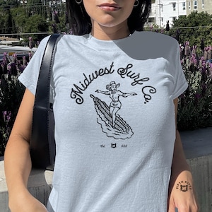 May include: A light blue short-sleeve t-shirt featuring a black graphic print of a person surfing on a corn cob. The text reads Midwest Surf Co. with Est. and 028 on either side of a small cat icon. A black shoulder bag strap is visible on the side.