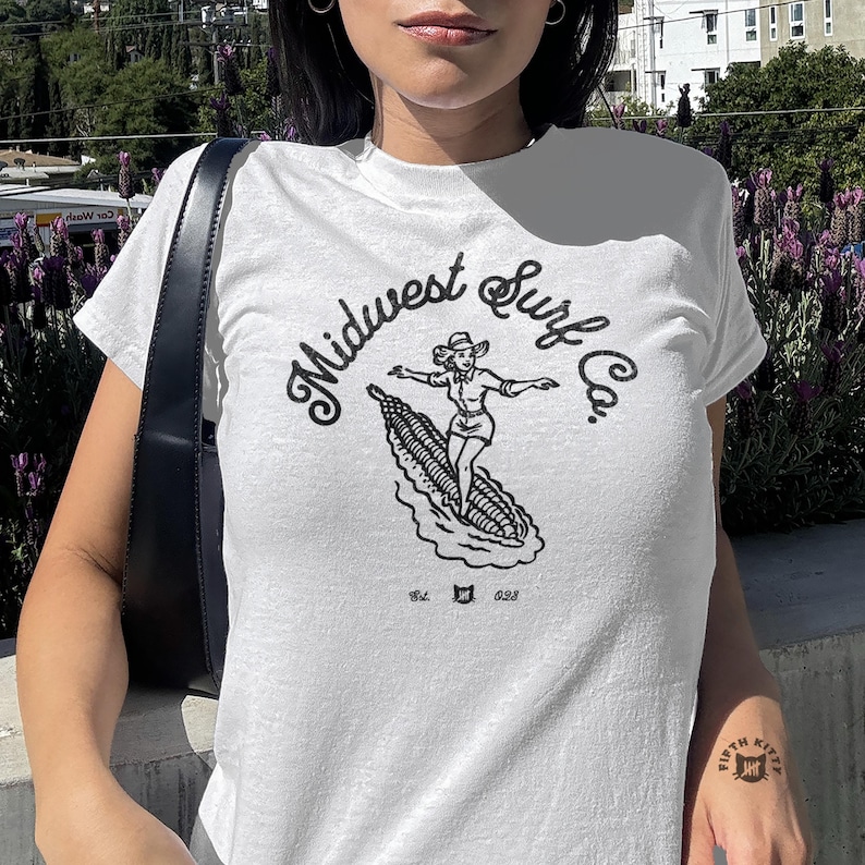 Vintage Midwest Surf Co. Baby Tee, Y2K Inspired Graphic Tee, 90s Retro Baby T-Shirt, Funky Graphic Design Tee, Nostalgic Baby Tee. Image of a country girl surfing on a corn cob with text Midwest Surf Co
