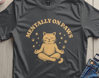 May include: Dark gray t-shirt with the text "MENTALLY ON PAWS" in a circular design. The shirt features a cartoon cat meditating in a lotus position, surrounded by stars and paw prints.