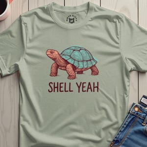 Könnte beinhalten: Hellgrünes T-Shirt mit einer detaillierten Illustration einer Schildkröte. Die Schildkröte hat einen türkisblauen Panzer und einen rötlich-braunen Körper. Der Text "SHELL YEAH" ist darunter in passendem Rotbraun gedruckt. Das Shirt hat ein Fifth Kitty-Logo am Halsausschnitt.
