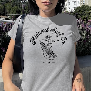 Vintage Midwest Surf Co. Baby Tee, Y2K Inspired Graphic Tee, 90s Retro Baby T-Shirt, Funky Graphic Design Tee, Nostalgic Baby Tee. Image of a country girl surfing on a corn cob with text Midwest Surf Co