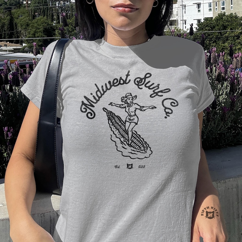 Vintage Midwest Surf Co. Baby Tee, Y2K Inspired Graphic Tee, 90s Retro Baby T-Shirt, Funky Graphic Design Tee, Nostalgic Baby Tee. Image of a country girl surfing on a corn cob with text Midwest Surf Co