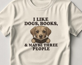 Può includere: T-shirt color crema con la scritta "I LIKE DOGS, BOOKS, & MAYBE THREE PEOPLE." Un cane marrone legge un libro al centro della maglietta. La maglietta è realizzata in un materiale morbido.