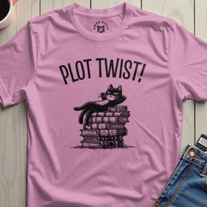 Puede incluir: Camiseta rosa claro con la frase "PLOT TWIST!" sobre un gato negro tumbado sobre una pila de libros. El gato tiene una expresión relajada. La camiseta tiene el logo de Fifth Kitty en el cuello.