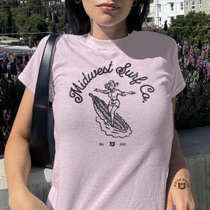 Vintage Midwest Surf Co. Baby Tee, Y2K Inspired Graphic Tee, 90s Retro Baby T-Shirt, Funky Graphic Design Tee, Nostalgic Baby Tee. Image of a country girl surfing on a corn cob with text Midwest Surf Co