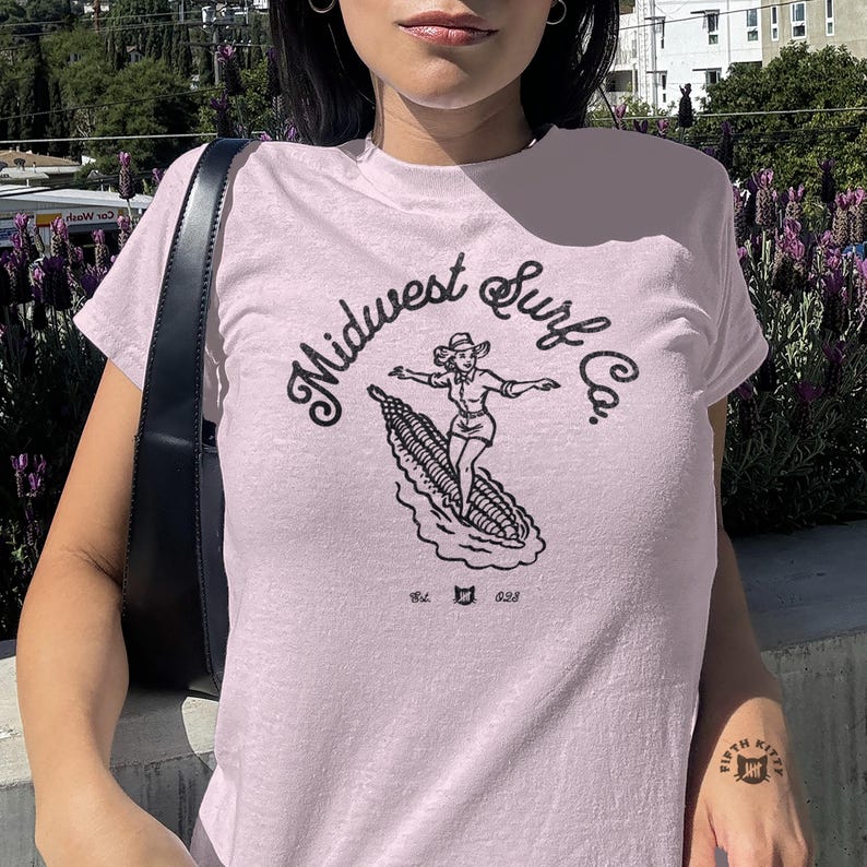 Vintage Midwest Surf Co. Baby Tee, Y2K Inspired Graphic Tee, 90s Retro Baby T-Shirt, Funky Graphic Design Tee, Nostalgic Baby Tee. Image of a country girl surfing on a corn cob with text Midwest Surf Co