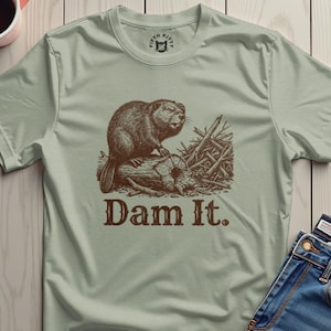 Könnte beinhalten: Salbeigrünes T-Shirt mit einer braunen Illustration eines Bibers auf einem Baumstamm und dem Text "Dam It." Der Biber ist von Ästen umgeben. Das T-Shirt hat einen Rundhalsausschnitt und kurze Ärmel.