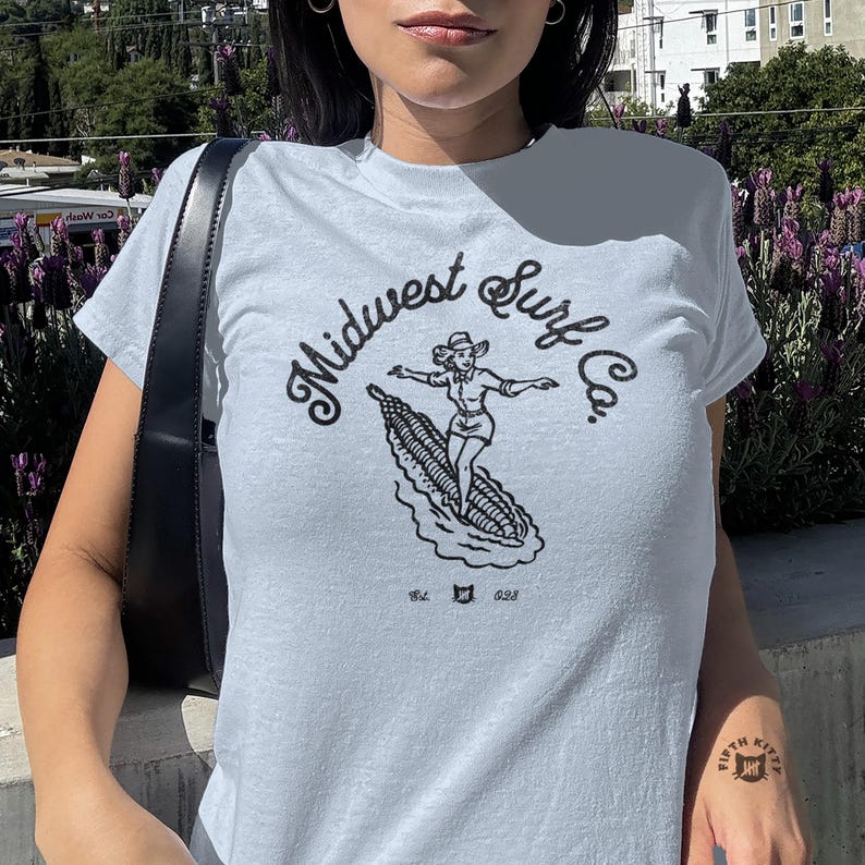 Może przedstawiać: Jasnoniebieski t-shirt z kr&oacute;tkim rękawem z czarnym nadrukiem graficznym przedstawiającym osobę surfującą na kolbie kukurydzy. Tekst brzmi Midwest Surf Co. z dopiskiem Est. i 028 po obu stronach małej ikony kota.