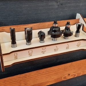 Puede incluir: Un estante de madera blanco con ocho ranuras para guardar fresas de router. El estante tiene etiquetas para cada ranura, incluyendo "Ø12 L20", "Ø12.7 R6", "Ø21.8 R6.3", "Ø25 R4.5" y "Ø12.7 1/4".