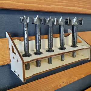 May include: A white wooden tool holder with six slots for holding drill bits. The holder is mounted on a wooden wall. The drill bits are black and have the Bosch brand name printed on them.