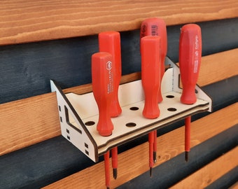 May include: A wooden screwdriver holder with a white frame and a dark brown trim, holding several red screwdrivers. The holder is mounted on a wooden wall with alternating dark and light brown planks. The screwdrivers are of varying sizes.