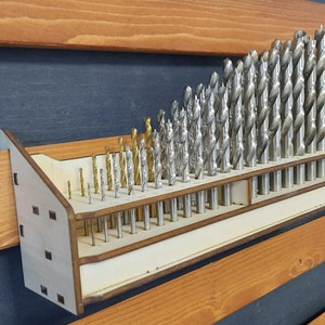 Może przedstawiać: Drewniany organizer na wiertła z jasnobrązowym wykończeniem. Organizer ma dwa rzędy otworów na wiertła. Organizer jest zamontowany na ścianie z ciemnoniebieskim i jasnobrązowym paskowanym tłem. Wiertła są ułożone według rozmiaru, od najmniejszych na górze do największych na dole.