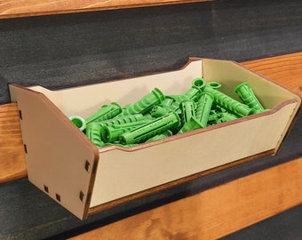 May include: A wooden wall-mounted storage bin with a beige finish holds a collection of green plastic wall anchors.