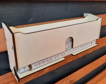 May include: White wooden wall-mounted organizer with a measuring tape inside. The organizer has a slot for the tape to slide through and a lip to hold it in place.