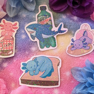 May include: Four axolotl stickers with glitter accents. One axolotl is in a jar labeled "Jam", another is on a slice of toast, another is in a shell, and the last is on top of a green Mountain Dew bottle.