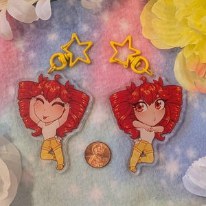 Puede incluir: Dos colgantes de acrílico transparente con una chica de dibujos animados con pelo rojo, que lleva una camiseta blanca y pantalones cortos amarillos. Cada colgante tiene un llavero amarillo en forma de estrella. Una moneda de un centavo se coloca entre los colgantes.