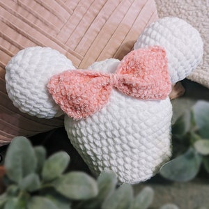 May include: A white crocheted plush toy shaped like a mouse with a pink velvet bow. The toy is sitting on a pink and white striped pillow.